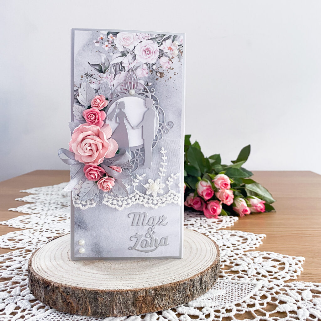 Ręcznie wykonana kartka ślubna w formacie DL. Cała w szaro srebrno różowej kolorystyce. Ozdobiona papierowymi różami, koronką i perełkami.