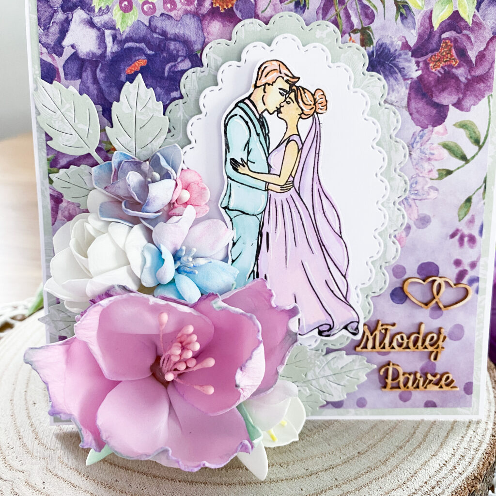 ręcznie wykonana kartka ślubna w fioletowej kolorystyce. Na kartce znajduje się bardzo rozbudowana kompozycja kwiatowa. Całość zapakowana jest w ozdobne pudełko.